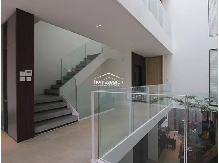This image captures a bright, modern atrium-style landing area featuring a striking staircase with glass balustrades and stone-tread steps. The space is finished with neutral large-format floor tiles and crisp white walls, creating an open, airy flow that connects different levels of the home. Architectural elements like the recessed ceiling lighting and the visible indoor greenery add a sophisticated, well-maintained touch to the interior.