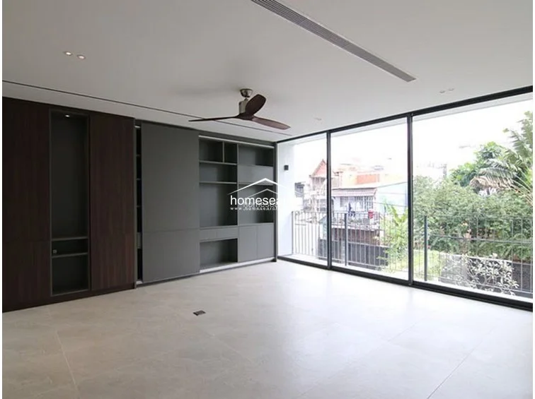 This spacious room features floor-to-ceiling glass sliding doors that provide abundant natural light and a view of the surrounding greenery. It is equipped with a modern ceiling fan, recessed lighting, and extensive custom built-in cabinetry in dark wood and grey tones, offering significant storage. The large-format light grey floor tiles and minimalist aesthetic suggest a high level of maintenance and contemporary design.