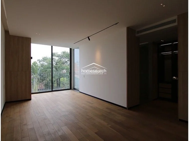 This bedroom or lounge area features warm wood-plank flooring and clean white walls, creating a cozy yet modern atmosphere. The room includes a large window with a balcony view, a sleek black track lighting fixture, and a walk-in wardrobe area tucked to the side. The space is in excellent condition, showcasing a thoughtful layout that maximizes natural light and provides a seamless transition to the dressing area.