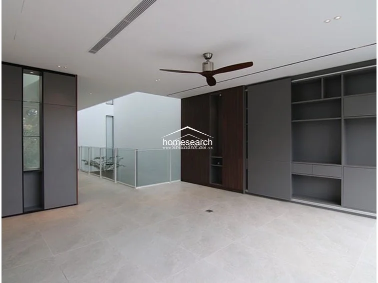 This wide-angle view highlights the open-plan layout of the upper level, emphasizing the connection between the landing and the adjacent living space. The design utilizes a consistent palette of light grey floor tiles, white walls, and dark wood accents, complemented by glass railings that maintain an unobstructed visual flow. The presence of a modern ceiling fan and integrated storage units confirms a high standard of functional, contemporary architecture.