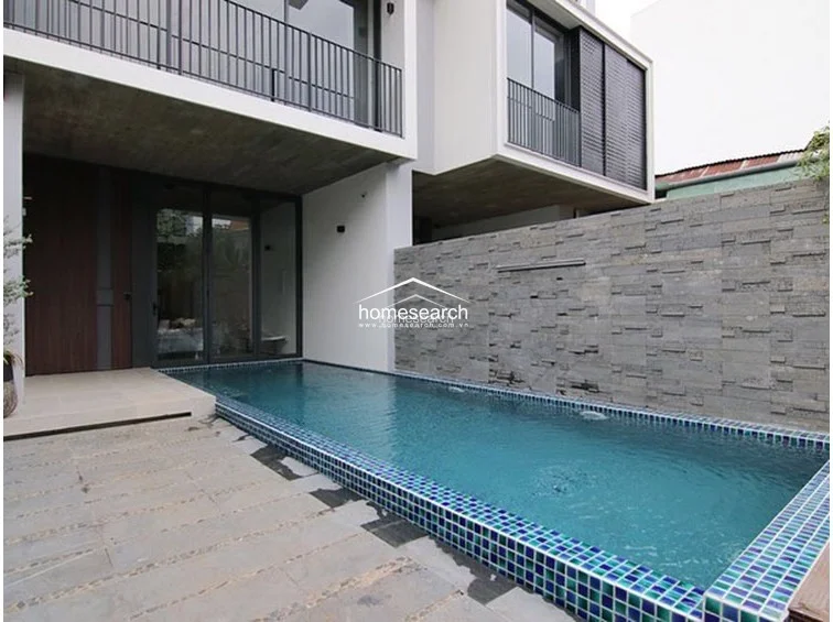 This exterior view showcases a modern, minimalist private swimming pool lined with vibrant blue tiles, set against a striking textured grey stone feature wall. The architecture features clean lines, a concrete overhang, and large floor-to-ceiling glass doors that seamlessly connect the outdoor pool area to the interior living space.