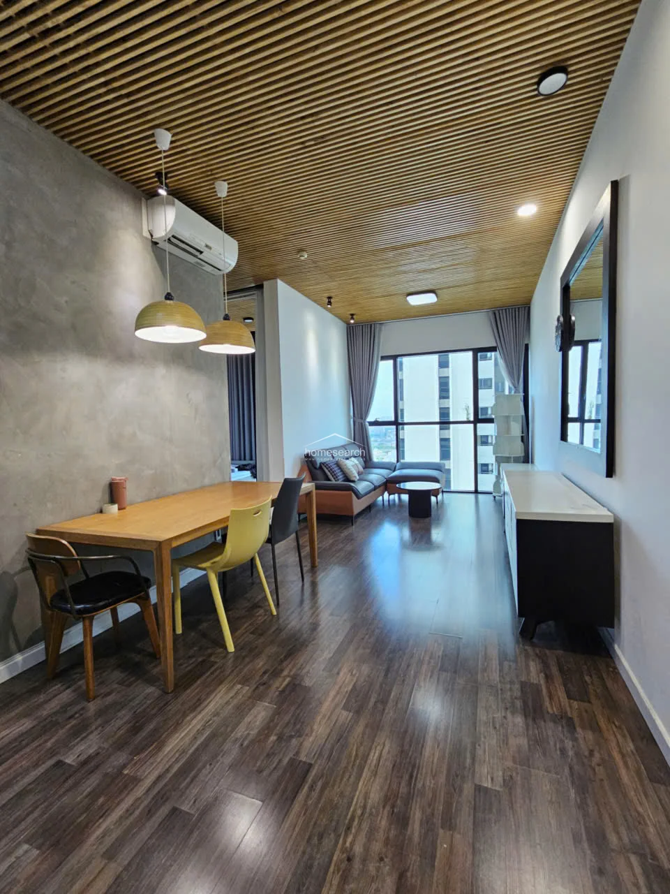 This open-concept living and dining area features a striking wood-slatted ceiling and dark wood-look flooring, creating a modern, warm atmosphere. The space is well-lit by natural light from the floor-to-ceiling windows and includes a wall-mounted AC unit, a concrete-textured accent wall, and a sleek dining set.