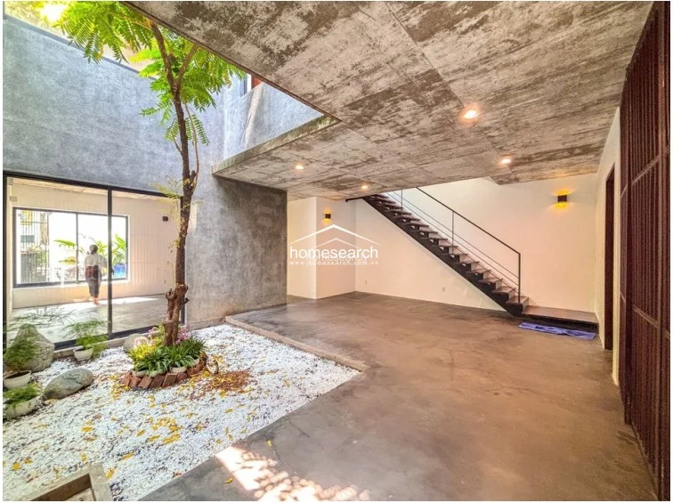 This interior courtyard space acts as a central light well, featuring raw concrete ceilings and polished concrete flooring that connects seamlessly to the adjacent living areas. A striking tree planted in a white gravel bed provides a natural focal point, while the open staircase with a minimalist metal railing adds to the industrial-chic aesthetic.