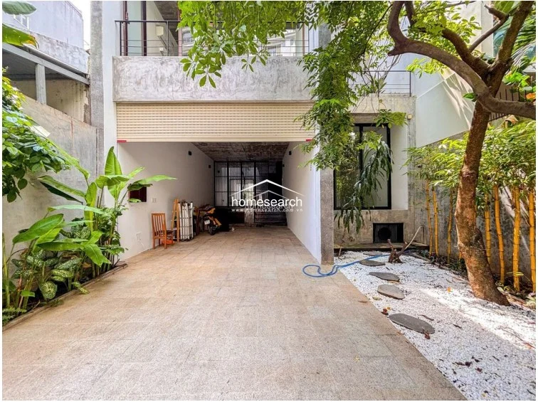 This exterior view showcases a modern, minimalist townhouse with a stone-paved driveway and a unique open-air courtyard featuring a mature tree and white gravel. The facade incorporates raw concrete finishes and a retractable security shutter, leading to a semi-open ground floor layout that emphasizes indoor-outdoor living.