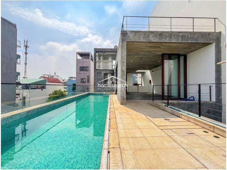 This outdoor terrace features a sleek, long lap pool with mosaic tiling and a glass railing that offers an unobstructed urban view. The adjacent covered patio area showcases a modern industrial aesthetic with a raw concrete ceiling and large sliding glass doors that provide seamless indoor-outdoor flow. The space is finished with durable stone tile flooring and includes a metal-railed balcony above, highlighting a well-maintained, contemporary architectural design.