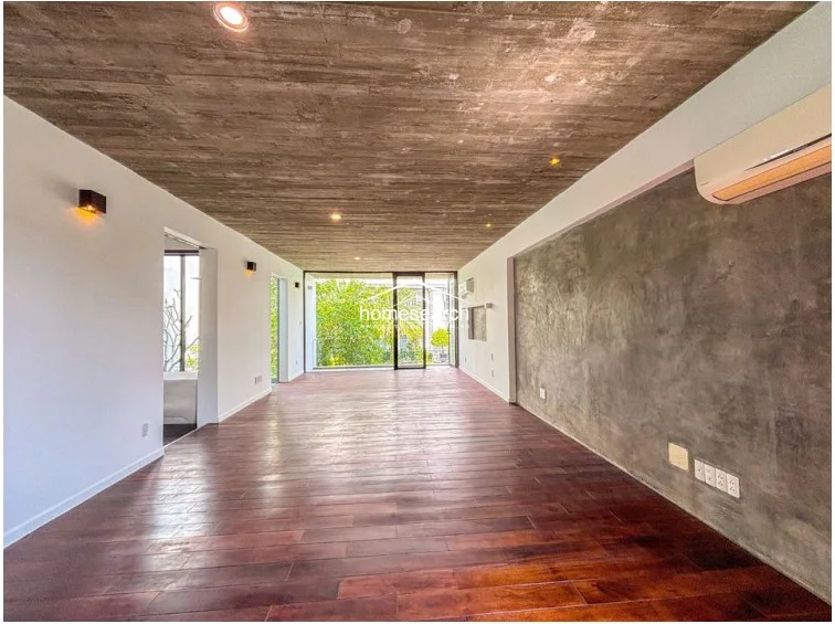 This spacious room features warm-toned wood-look flooring and a raw concrete ceiling, complemented by a large feature wall in a matching concrete finish. The space is equipped with a wall-mounted AC unit and recessed lighting, offering a bright, airy environment with direct access to a balcony overlooking lush greenery.
