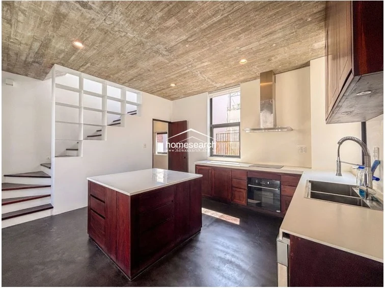 The kitchen features a sophisticated design with a large dark wood island, white countertops, and integrated appliances including an oven and range hood. The space is defined by a raw concrete ceiling and dark flooring, with built-in white shelving under the staircase providing clever, integrated storage solutions.