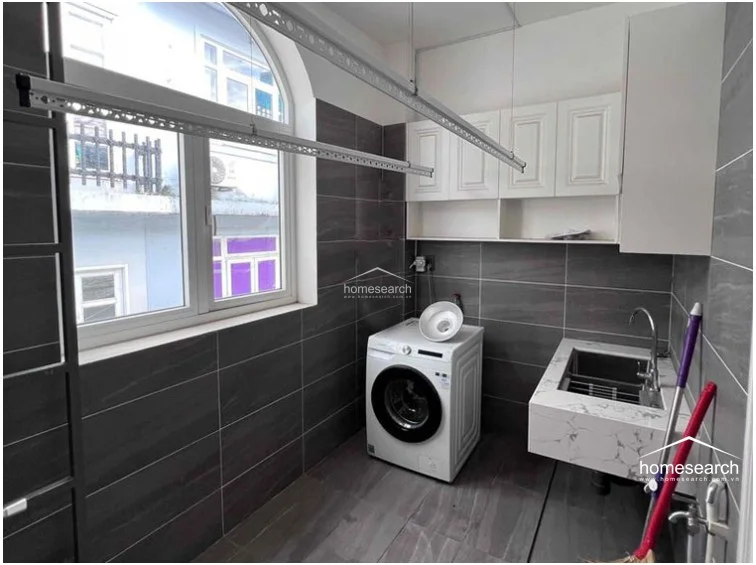 This functional laundry room or utility area is finished with the same dark grey tiling as the bathroom, creating a cohesive design theme throughout the home. It is equipped with a front-loading washing machine, a white marble-topped utility sink, and ample white cabinetry for storage. The space benefits from a large, arched window that provides significant natural light and includes a ceiling-mounted retractable drying rack for added convenience.