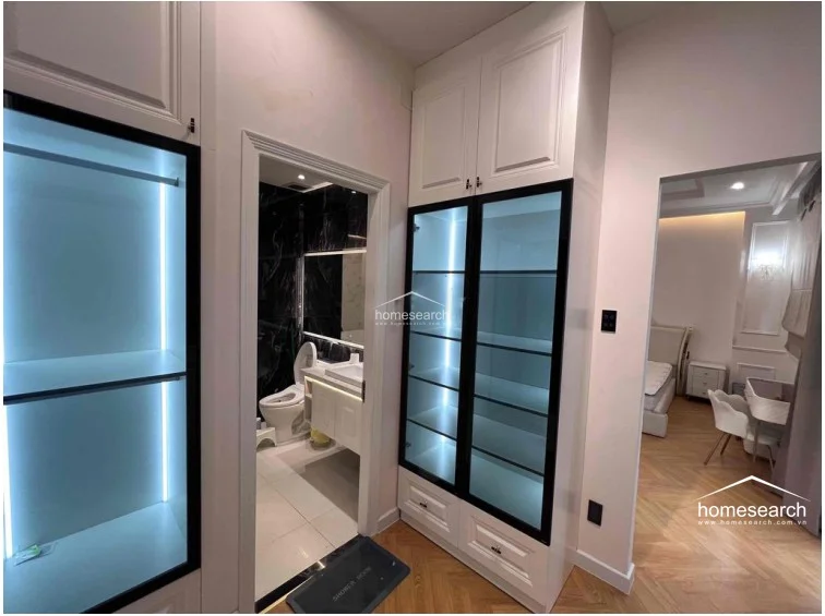 This dressing area serves as a transitional space, featuring custom floor-to-ceiling cabinetry with glass-fronted doors and integrated LED lighting. It provides a seamless connection between the bedroom and the adjacent bathroom, which is finished with high-contrast black marble-style wall tiles and white fixtures.