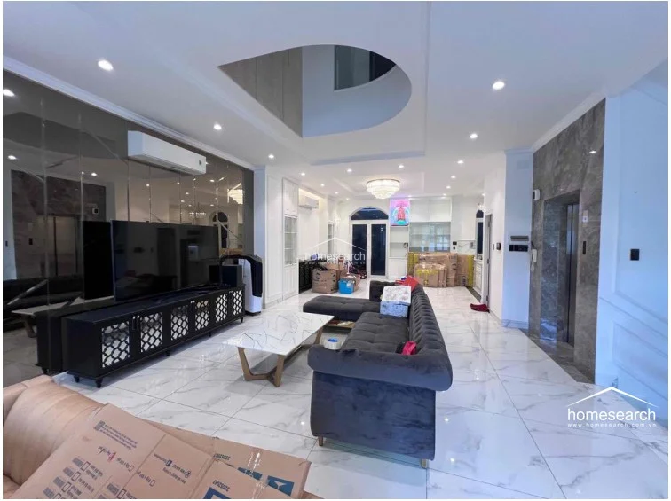 This spacious, open-plan living area is defined by luxurious white marble flooring and a high-gloss mirrored wall that amplifies the sense of space. The room is equipped with a modern wall-mounted AC unit, a sleek black media console, and a contemporary grey sectional sofa, all illuminated by a central crystal chandelier and recessed lighting.