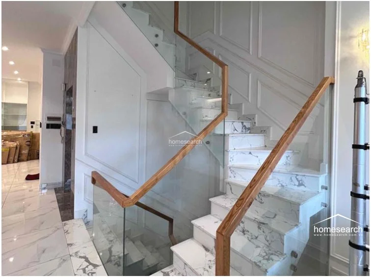 This elegant staircase serves as a central architectural feature, utilizing a modern combination of white marble steps with grey veining, clear glass railings, and warm-toned wooden handrails. The surrounding walls are adorned with classic wainscoting, and the design flows seamlessly into the adjacent living and kitchen spaces, maintaining a bright and airy aesthetic.
