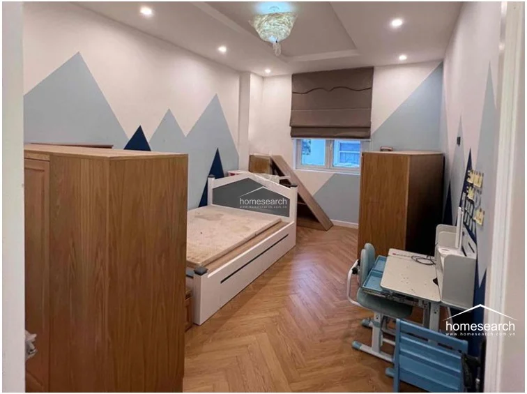 This children's bedroom features a playful mountain-themed wall mural in shades of blue, paired with warm, light-colored herringbone wood flooring. The room is furnished with a white platform bed, two large wooden wardrobes for ample storage, and a dedicated study area, all illuminated by recessed ceiling lights and a unique decorative pendant fixture.