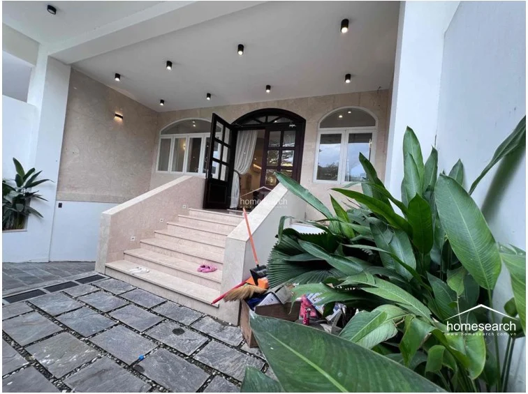 The entryway features a grand set of stone stairs leading to a sophisticated arched double-door entrance, flanked by lush greenery. The exterior walls are finished in a warm, neutral stone cladding, and the area is well-lit with modern recessed ceiling lights, creating a welcoming and upscale transition into the home.