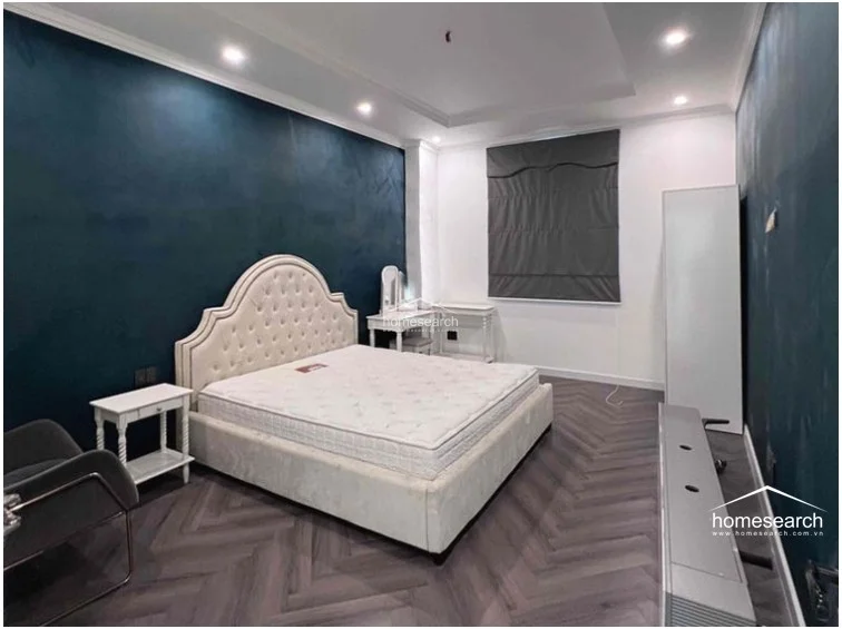 This bedroom features a sophisticated dark blue accent wall that contrasts with white walls and a light-colored, tufted upholstered bed frame. The room is finished with dark wood herringbone flooring, a minimalist white vanity, and a standalone wardrobe, all illuminated by recessed ceiling lights.