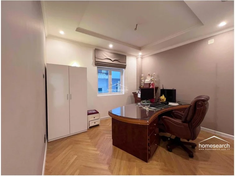 This functional home office is equipped with a large, dark wood executive desk and a matching leather office chair, set against a neutral, warm-toned color palette. The space includes a white wardrobe for storage and features elegant herringbone wood flooring, with natural light provided by a single window covered by a Roman shade.