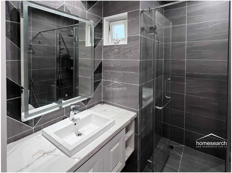 This modern bathroom features dark, large-format grey wall and floor tiles contrasted by a white marble-topped vanity and a sleek, LED-backlit mirror. It includes a glass-enclosed walk-in shower and a small, high-placed window that provides natural ventilation and light while maintaining privacy. The overall condition is excellent, showcasing a contemporary aesthetic with clean lines and high-quality fixtures.