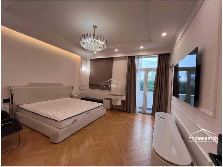This primary bedroom exudes luxury with a grand crystal chandelier, a plush upholstered bed, and a sleek white television console. The room flows directly onto a private balcony through glass doors, and the herringbone wood flooring is complemented by soft, neutral wall tones and recessed lighting.