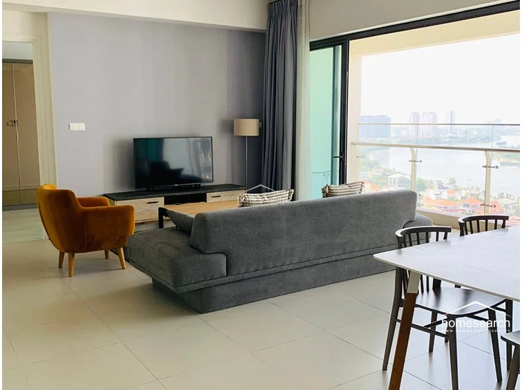This view emphasizes the transition from the living area to the outdoor balcony, which features a glass railing to ensure an unobstructed view of the river and cityscape. The room is decorated with a neutral color palette, including grey walls and matching curtains, which complements the contemporary furniture. The overall condition of the space is pristine, reflecting a high level of maintenance.