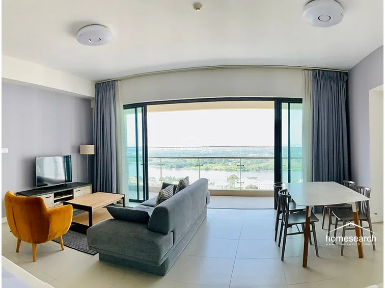 This open-concept living and dining area features a bright, modern layout with a seamless flow toward a large balcony offering panoramic river views. The space is finished with neutral light-colored floor tiles, soft grey walls, and floor-to-ceiling glass doors that maximize natural light. Furnishings include a contemporary grey sofa, a vibrant mustard-colored accent chair, and a minimalist dining set, all maintained in excellent condition.