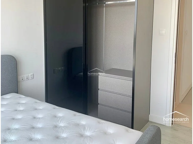 The bedroom includes a sleek, built-in wardrobe with dark, reflective sliding doors and integrated drawer storage. The minimalist design is complemented by light wood-tone flooring and a neutral color palette, maintaining a contemporary and organized feel.