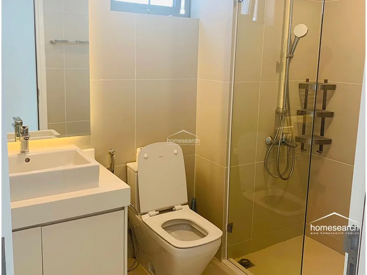 The bathroom is well-maintained, featuring a glass-enclosed walk-in shower, a modern vessel sink, and light-colored wall tiles. It includes essential fixtures like a handheld showerhead and a wall-mounted storage rack, offering a compact and functional layout.