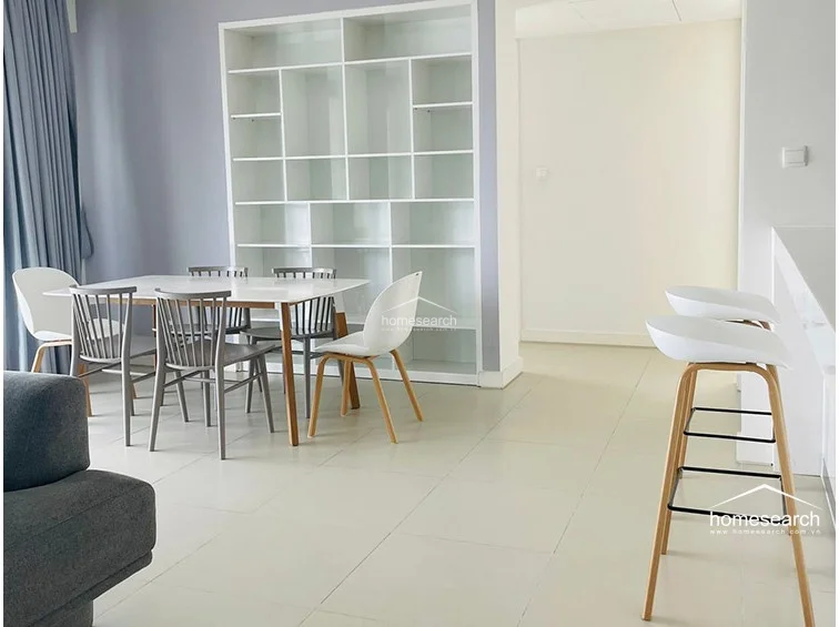 This perspective showcases the dining area and the adjacent kitchen breakfast bar, highlighting the efficient use of space in this open floor plan. The dining set is positioned near a floor-to-ceiling shelving unit, while the white breakfast bar stools provide additional seating and a clear visual boundary for the kitchen. The flooring consists of large, light-toned tiles that unify the entire living area.