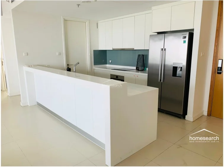 The kitchen features a sleek, modern design with white cabinetry, a clean white countertop, and a contrasting blue-grey backsplash. It is equipped with high-end stainless steel appliances, including a large side-by-side refrigerator and an integrated cooktop. The layout is functional and minimalist, maintaining the property's overall aesthetic of modern, well-kept luxury.