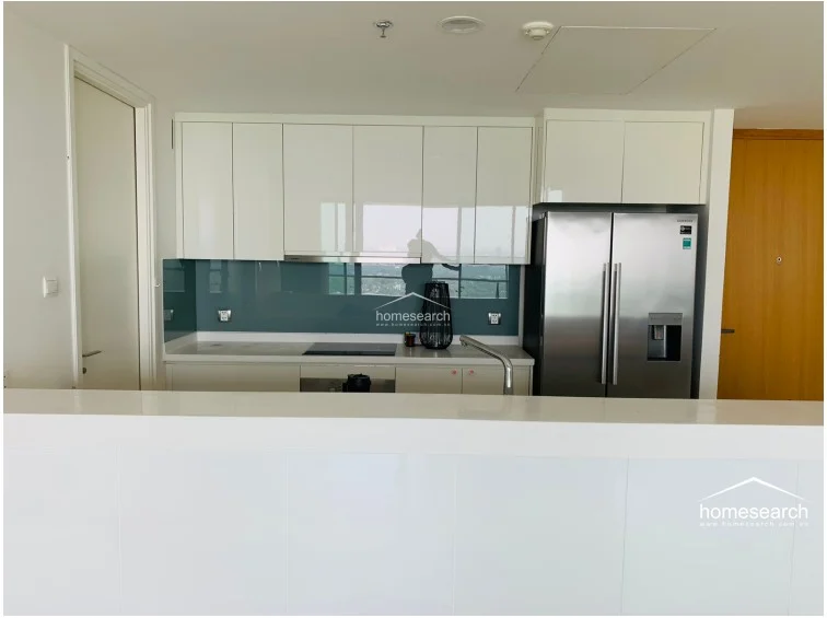 This modern kitchen features a clean, minimalist layout with high-gloss white cabinetry and a contrasting teal glass backsplash. It is equipped with a stainless steel double-door refrigerator and an integrated induction cooktop, all set against a neutral white countertop that serves as a breakfast bar.