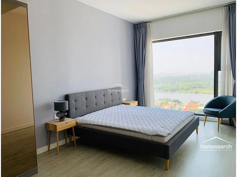 This bedroom offers a serene atmosphere with light wood-look flooring and soft grey walls, centered around a contemporary tufted grey headboard. A large floor-to-ceiling window provides ample natural light and a stunning panoramic view of the river, complemented by elegant floor-length curtains.