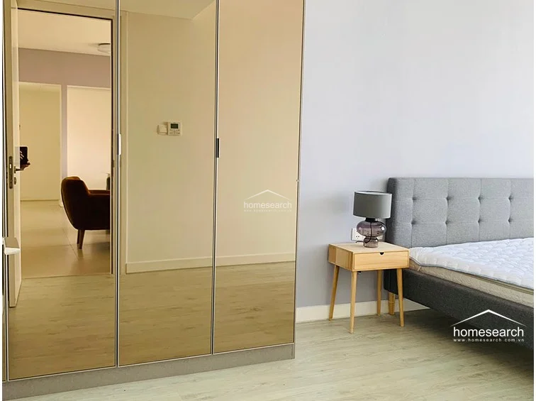 The bedroom includes a functional built-in wardrobe with mirrored doors that reflect the adjacent living space, enhancing the room's sense of depth. The minimalist decor is completed by light wooden bedside tables and a modern grey lamp, maintaining a cohesive and well-maintained aesthetic.