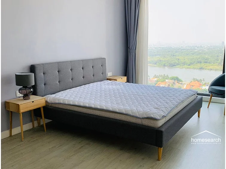 This bedroom focuses on the comfortable sleeping area, highlighting the quality of the grey upholstered bed frame and the light-toned wooden nightstands. The room is well-lit by natural light streaming through the expansive window, which frames a scenic river view.