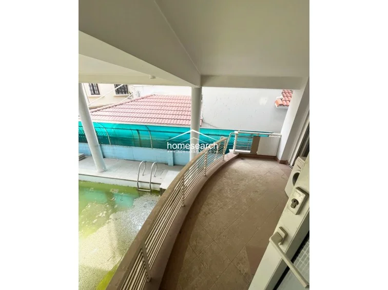 This elevated balcony overlooks the pool area and is finished with neutral-toned floor tiles and metal safety railings. While the balcony offers a good vantage point and potential for outdoor seating, the pool below remains in poor condition, indicating that the property requires comprehensive renovation and exterior care.