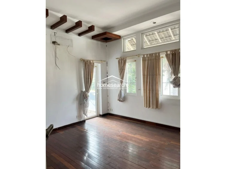 This living area features rich, dark hardwood flooring and white walls, accented by unique wooden ceiling beams and a box-style architectural detail. The space is filled with natural light from multiple windows and a glass-paneled door, while the exposed wiring suggests a need for minor electrical maintenance.
