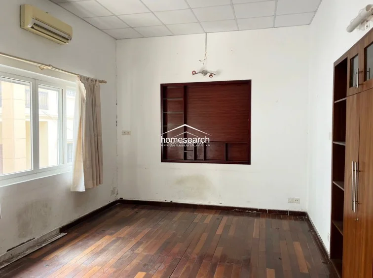This bedroom includes a built-in wooden wardrobe and a recessed shelving unit, providing functional storage against a clean white backdrop. The room is equipped with a wall-mounted AC unit and features durable dark hardwood flooring, though the walls show some signs of moisture staining near the floor.