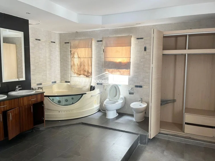 This expansive master bathroom is designed with a luxurious, spa-like layout, featuring a large corner jetted tub and a dedicated vanity area. It includes a built-in light wood wardrobe for storage, dark grey floor tiles, and textured wall tiles, offering a sophisticated and well-maintained environment.
