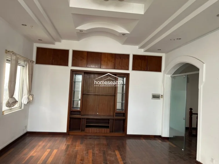 This living area features rich dark hardwood flooring and a prominent built-in wooden media console with overhead storage cabinets. The room is accented by a decorative tray ceiling with recessed lighting and connects to an adjacent space through a stylish arched doorway fitted with a frosted glass door.