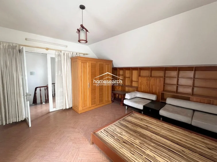 This attic-style bedroom features a sloped ceiling and a large, integrated wooden shelving unit that spans the wall. The room is furnished with a wooden bed frame and a matching wardrobe, with light-toned herringbone-patterned flooring and a unique hanging pendant light.