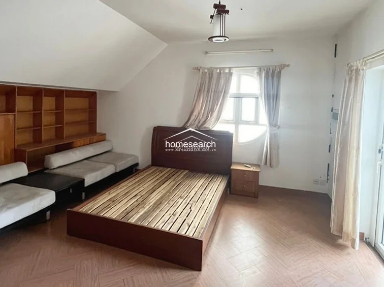 This bedroom layout utilizes the sloped roofline to create a cozy nook, featuring a circular window that adds an interesting architectural touch. The room is equipped with a wooden bed, a nightstand, and a built-in shelving system, all set against a neutral color palette with herringbone-style flooring.