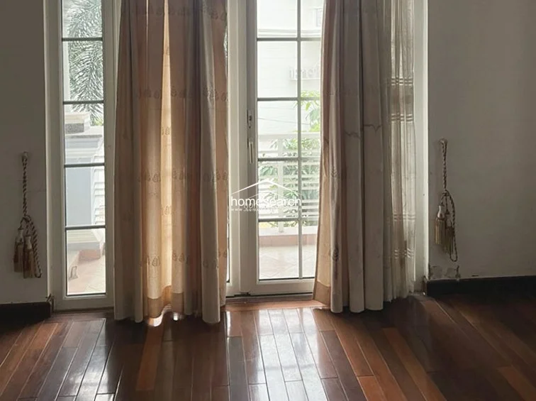 This bedroom or living area features rich, warm-toned hardwood flooring and floor-to-ceiling glass doors that open onto a private balcony. The space is filled with soft natural light filtered through elegant, neutral-colored curtains, providing a serene and inviting atmosphere.