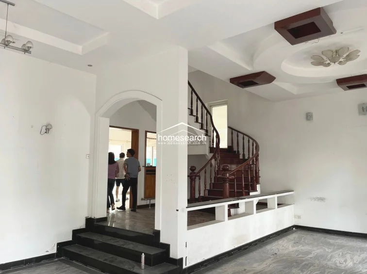 This spacious living area features a grand, curved wooden staircase as a central architectural focal point, complemented by a decorative arched entryway leading to an adjacent room. The space is finished with white walls, recessed ceiling details, and a polished marble floor, creating an open and airy flow.