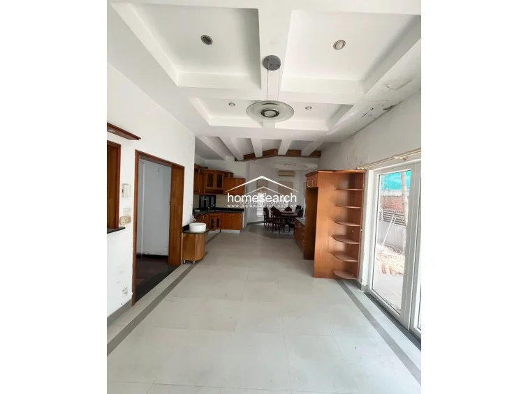 This large, open-plan kitchen and dining area is anchored by a functional L-shaped wooden cabinetry layout with dark countertops and a neutral tiled floor. The space flows seamlessly toward a bright dining nook surrounded by windows, and it includes a dedicated tall storage unit and a ceiling-mounted light fixture.