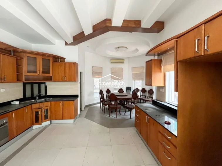 This expansive kitchen is fully equipped with extensive wooden cabinetry, dark stone countertops, and integrated appliances, offering a warm and traditional aesthetic. The dining area is positioned in a curved alcove with multiple windows, providing ample natural light and a view of the outdoors, all tied together by a unique ceiling design with wooden beams.