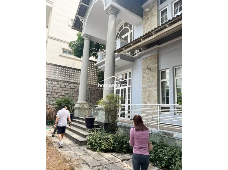 This exterior view showcases a multi-story home featuring grand architectural columns and a combination of light blue painted walls with natural stone accents. The property includes a paved stone walkway leading to a raised porch area, though the overall condition suggests the exterior paint and landscaping require significant maintenance.