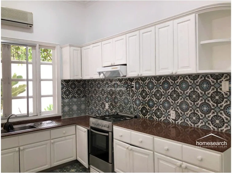 This kitchen features a functional L-shaped layout with classic white shaker-style cabinetry and striking patterned backsplash tiles that provide a bold focal point. The space is equipped with a stainless steel gas range, a range hood, and a deep sink, all set against a durable dark granite countertop. Natural light is provided by a large multi-pane window, and the room includes a wall-mounted AC unit for climate control.