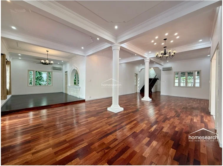 This expansive, open-plan living area features rich, polished hardwood flooring and high ceilings accented by elegant crown molding and structural columns. The space is flooded with natural light through multiple windows and is illuminated by sophisticated chandeliers, while a raised platform area adds architectural interest and defines the flow between zones. The clean white walls and neutral color scheme provide a bright, airy atmosphere, complemented by wall-mounted AC units for comfort.