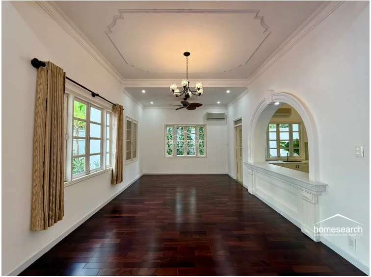 This elegant living area is defined by a beautiful arched pass-through window that connects to the kitchen, creating an open and airy atmosphere. The room features dark hardwood flooring, crisp white walls, and a classic chandelier, offering a sophisticated space for entertaining. Large windows provide ample natural light, and the inclusion of an AC unit ensures comfort in this well-maintained interior.