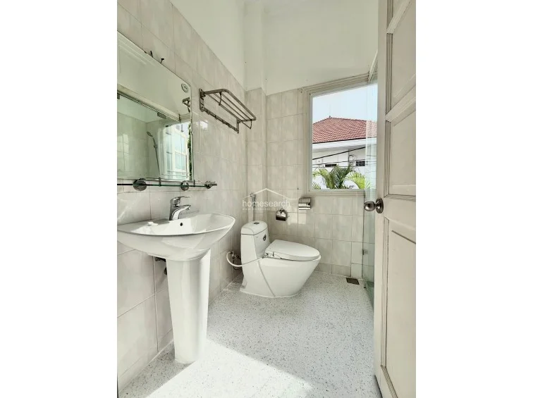 This compact, well-maintained bathroom features a pedestal sink, a modern toilet, and a glass-enclosed shower area. The space is finished with light-toned wall tiles and speckled flooring, with a window providing a view of the surrounding neighborhood.