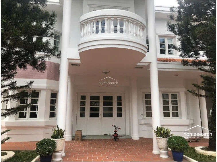 This striking exterior view showcases a grand, colonial-inspired architectural style with a prominent white facade, tall columns, and a decorative balcony. The entrance is framed by a terracotta-tiled porch and lush greenery, creating a welcoming and sophisticated curb appeal. The property appears to be in excellent condition, reflecting a high level of maintenance and classic design.