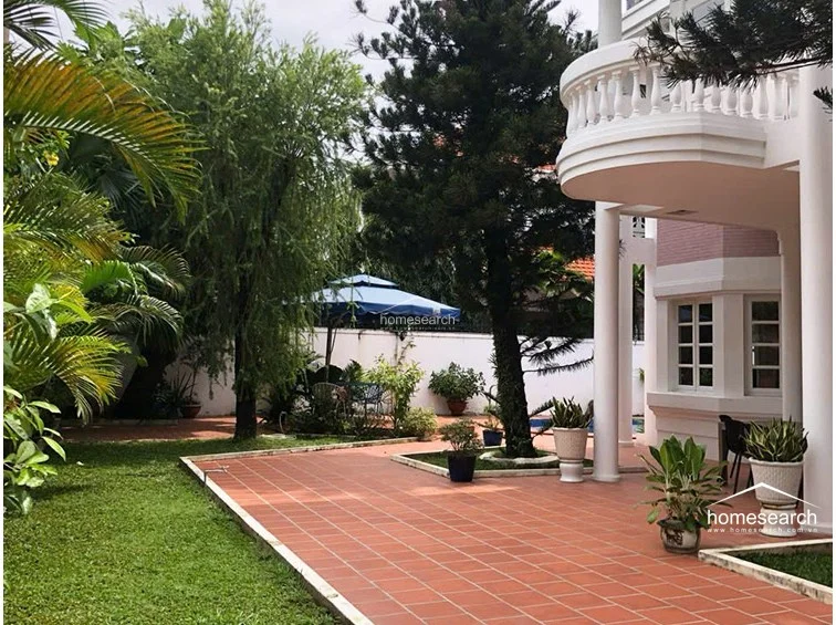 This expansive outdoor patio is paved with terracotta tiles and shaded by large, mature trees, providing a perfect setting for al fresco dining or lounging. The area is well-maintained and connects seamlessly to the main house, which features a distinctive curved balcony with white balustrades.