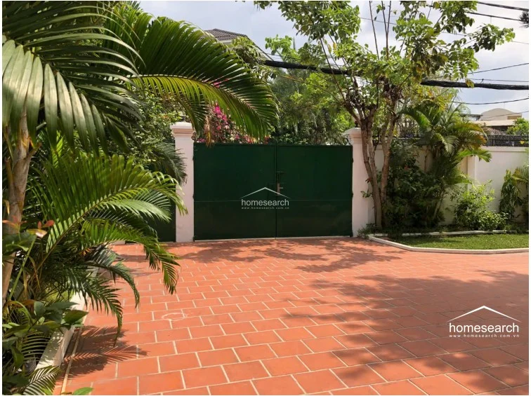 This entrance area features a secure, solid green metal gate set against white stucco pillars, providing both privacy and a classic look. The driveway is paved with durable terracotta tiles and is framed by vibrant tropical foliage and a small, well-kept lawn.